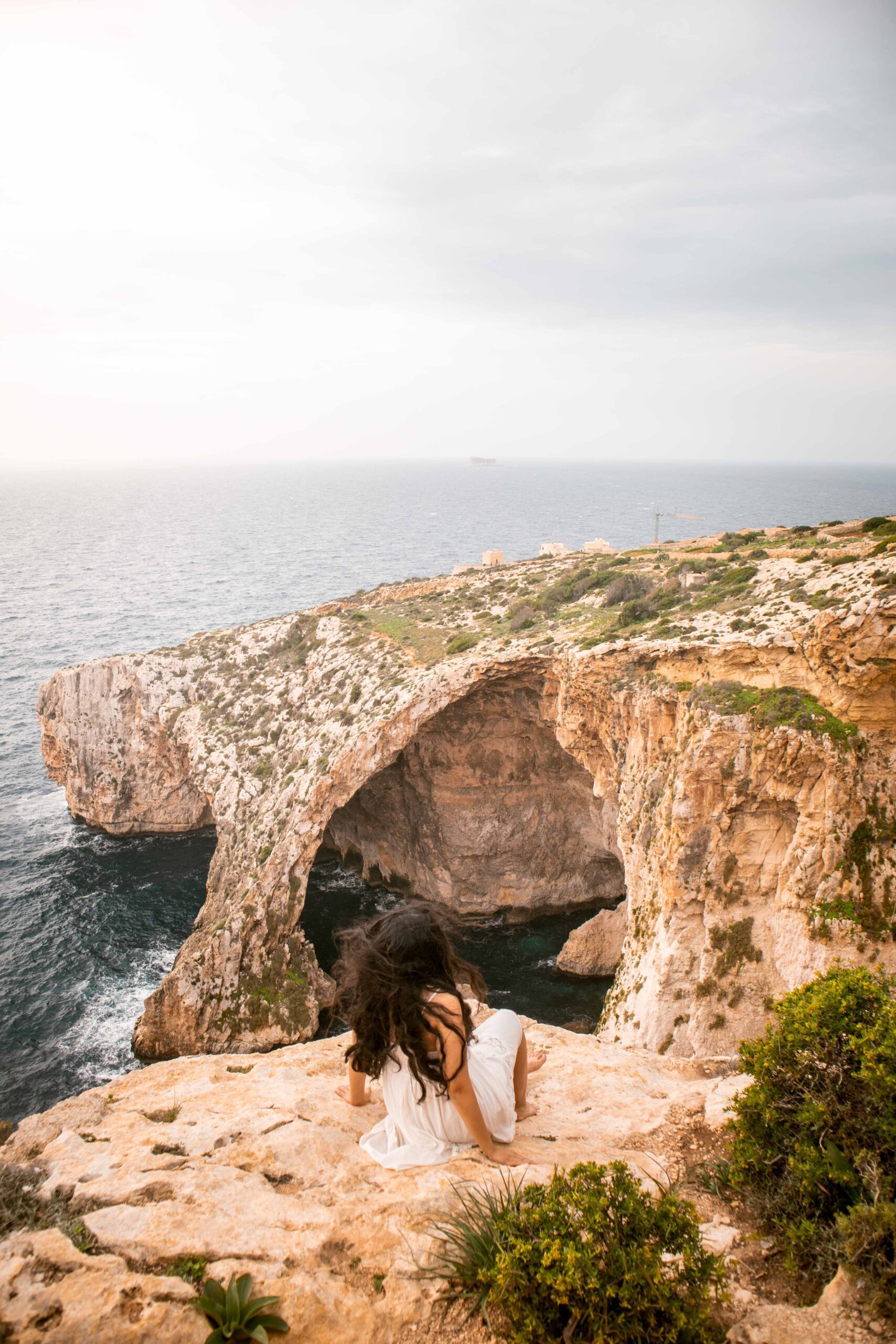 Woman wearing a white dress sitting on a cliff to watch the Blue Grotto from a Viewpoint in Wied Iż-Żurrieq, Malta