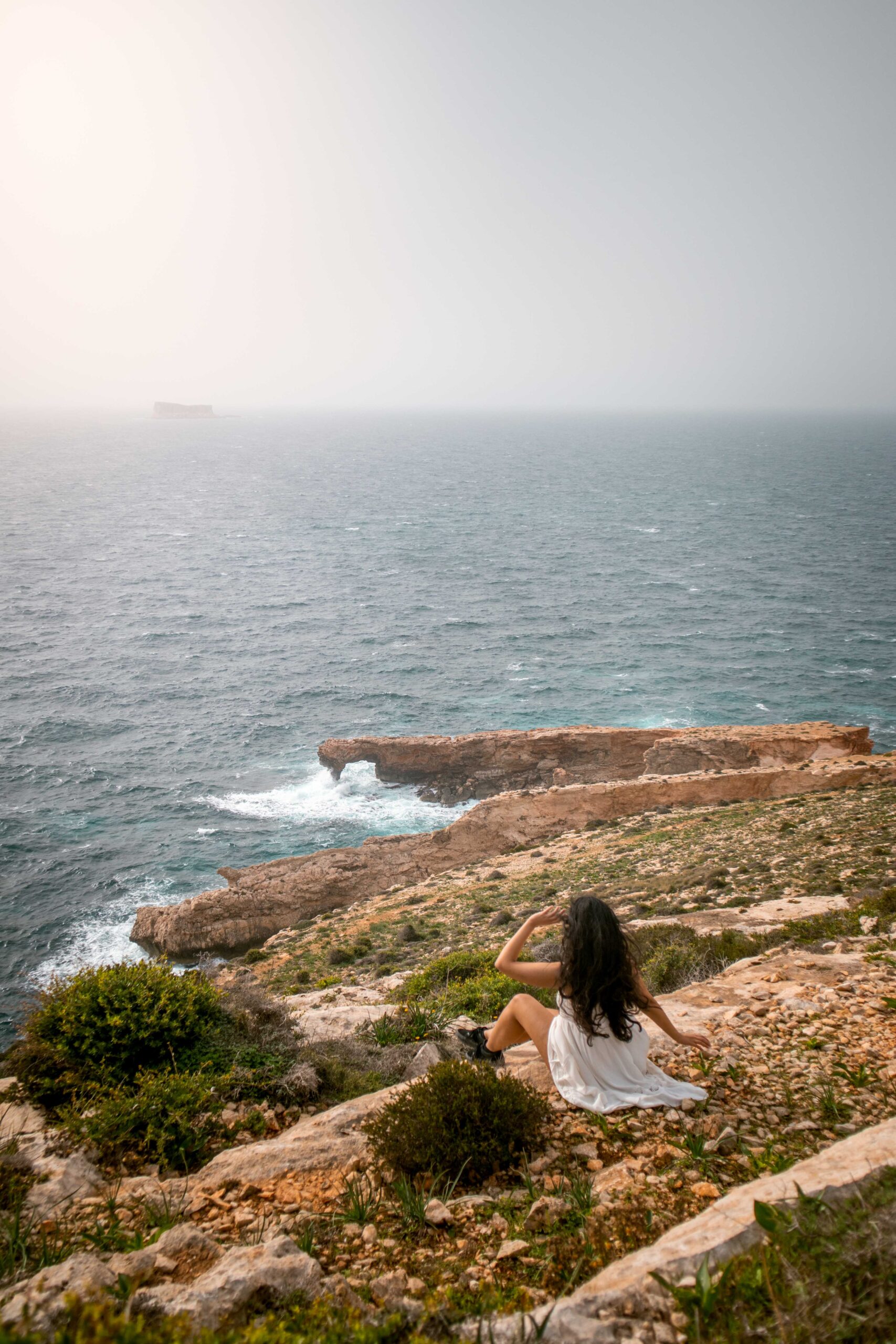 Woman wearing a white dress sitting and watching Għar Ħanex window near Ħaġar Qim and Mnajdra archaeological park in Il-Qrendi, Malta