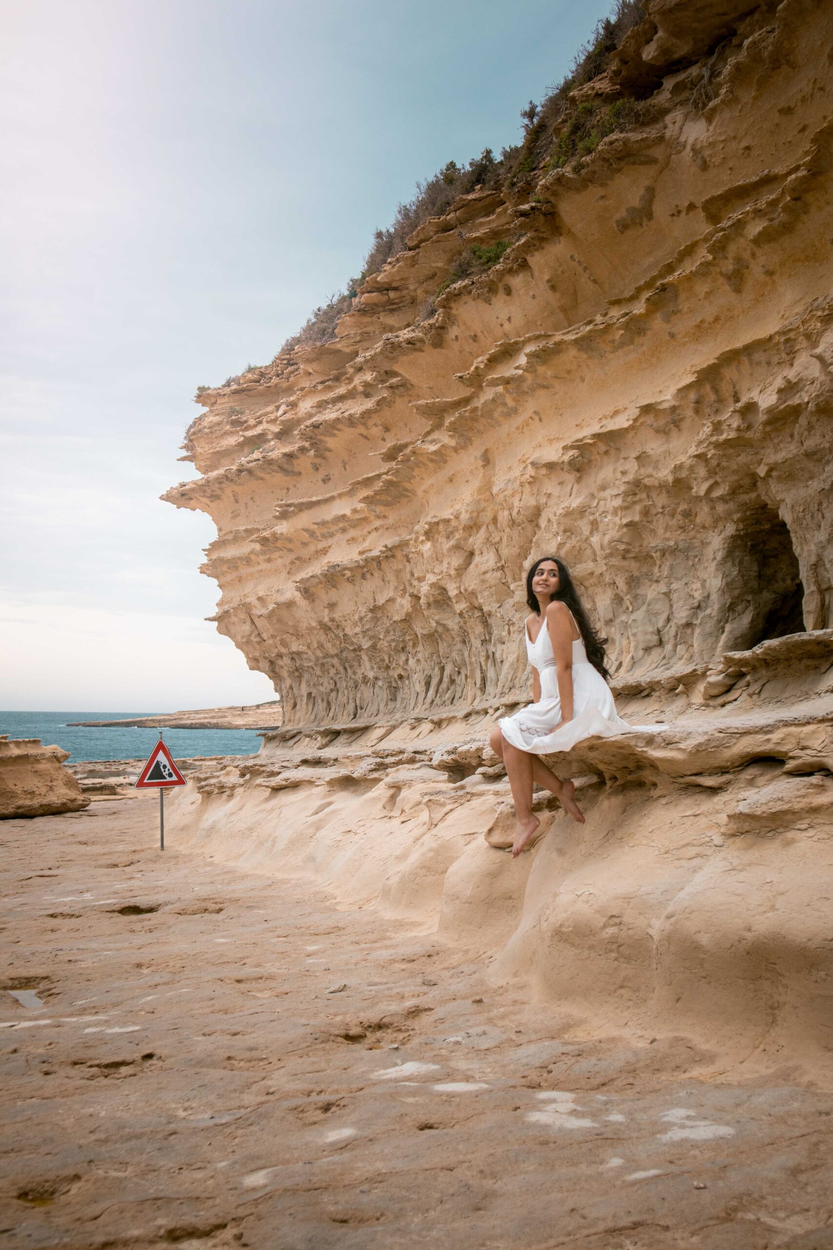 Woman wearing a white dress sitting near the cliffs of the Il-Qali hiking area located near Saint Peter's Pool in Malta