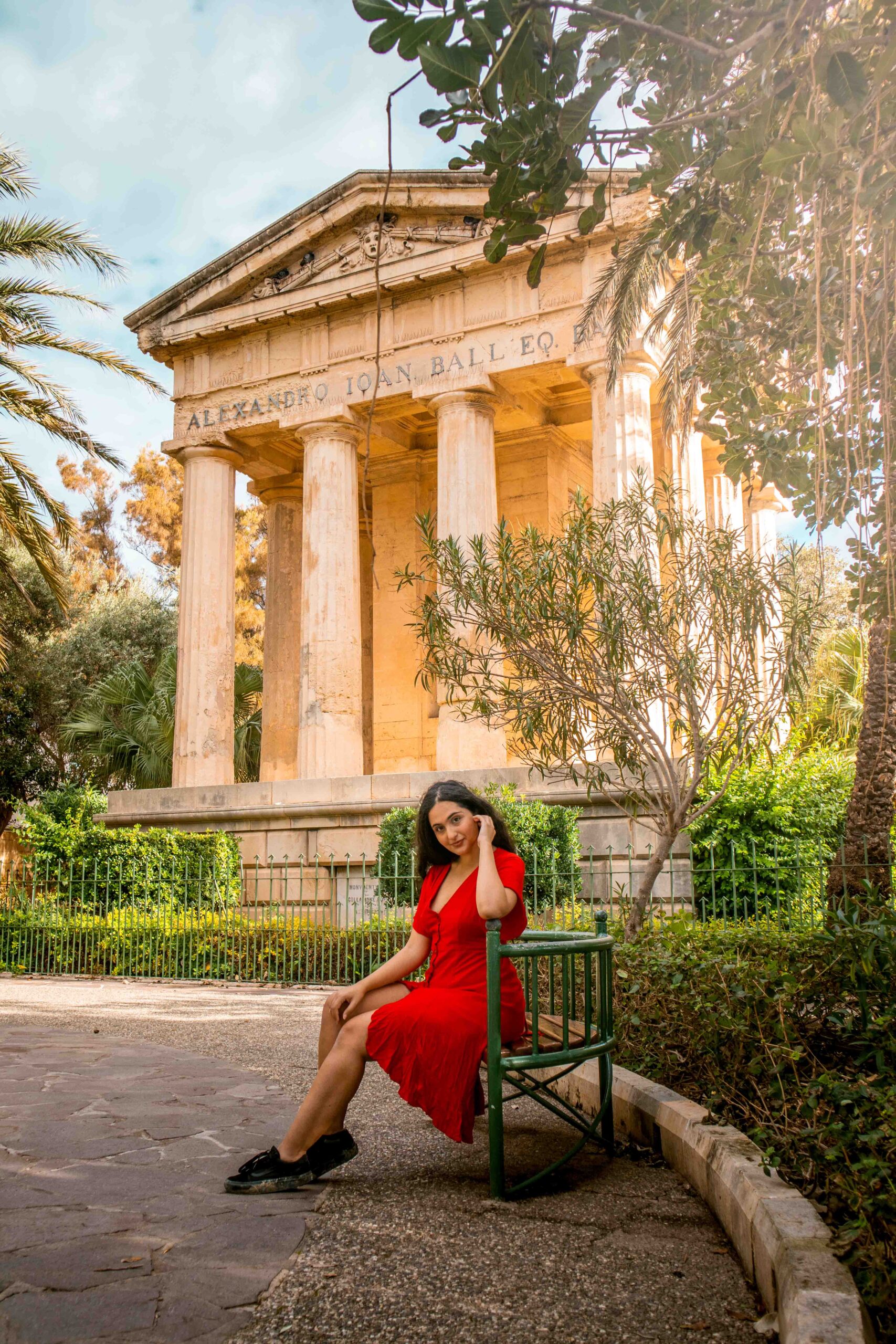 Woman wearing a red dress sitting on a bench in front of the Monument to Sir Alexander Ball in the Lower Barrakka Gardens in Valletta, Malta