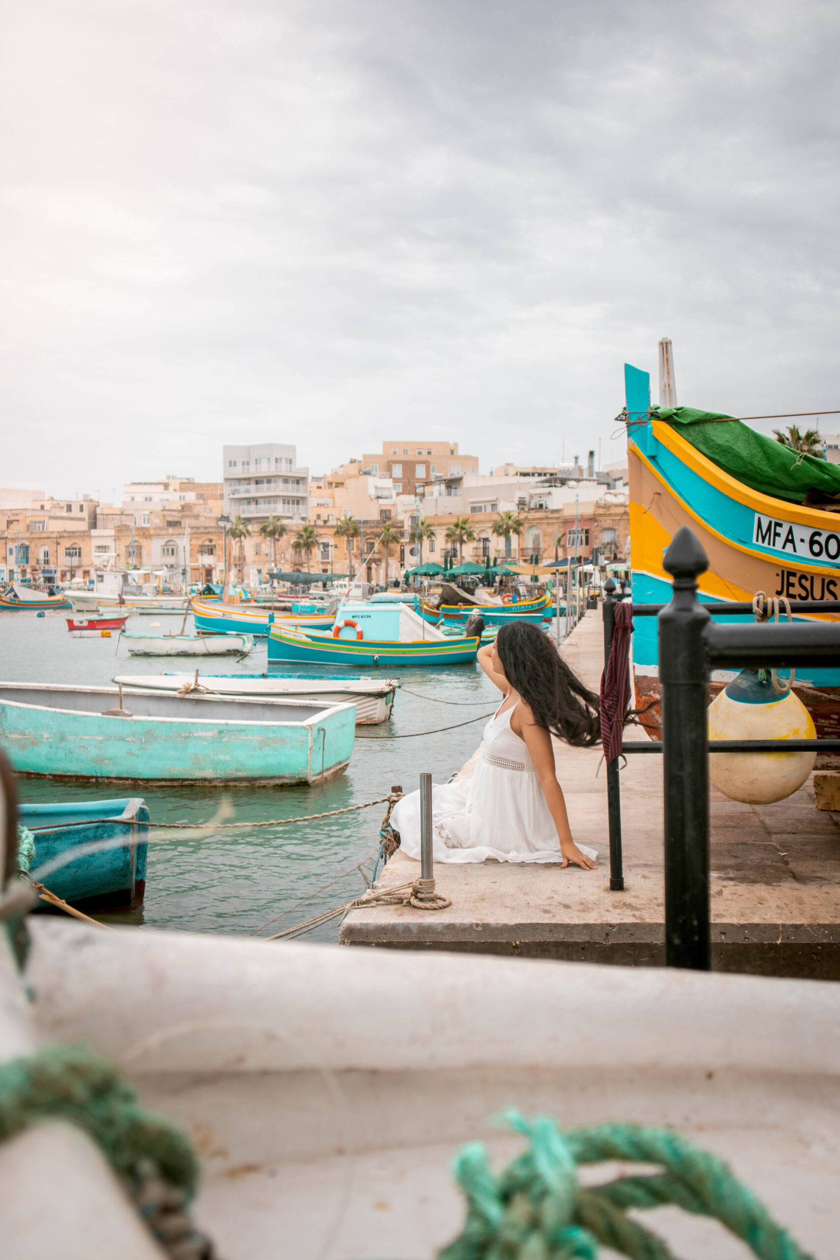 Woman wearing a white dress sitting in Marsaxlokk Harbour, Malta watching the waterfront on traditional colourful luzzu boats