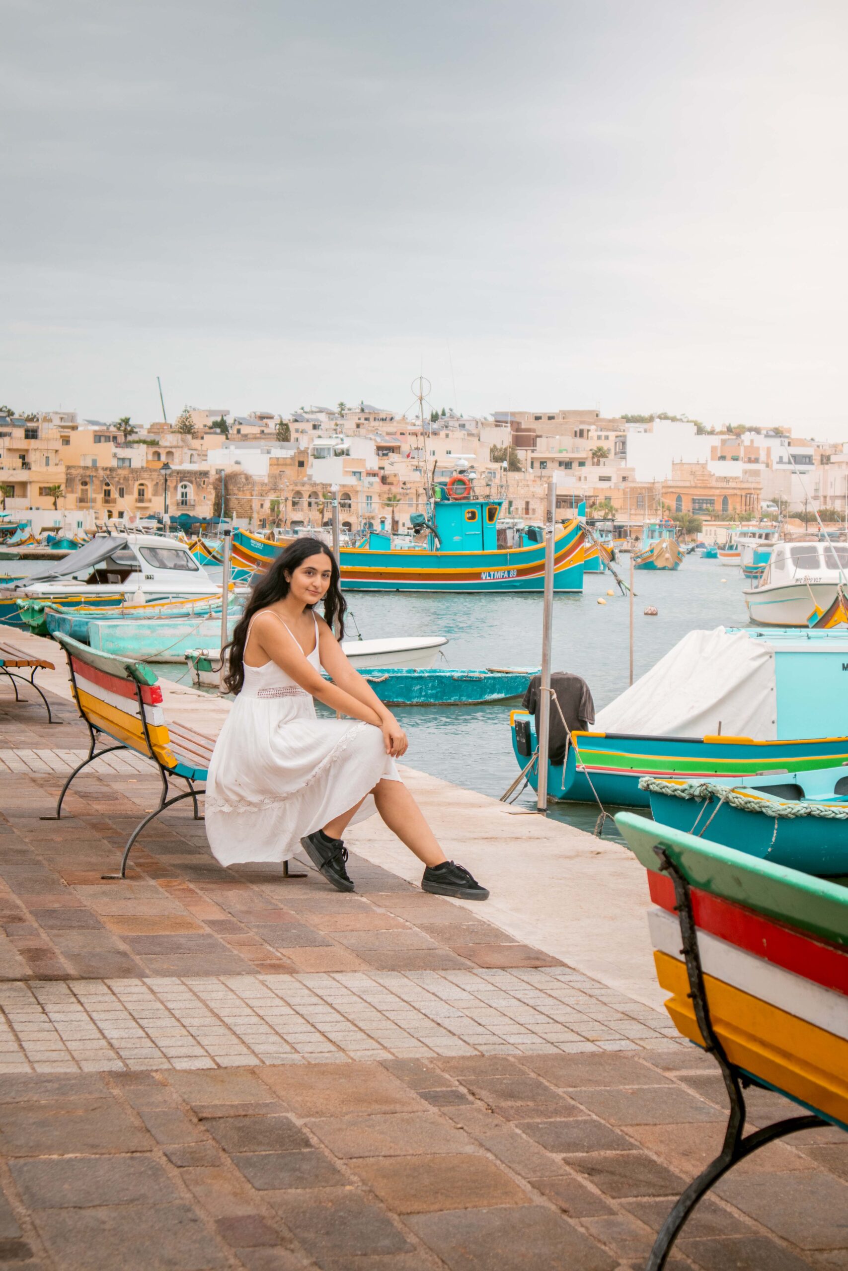Woman wearing a white dress sitting on a colourful bench in Marsaxlokk Harbour, Malta