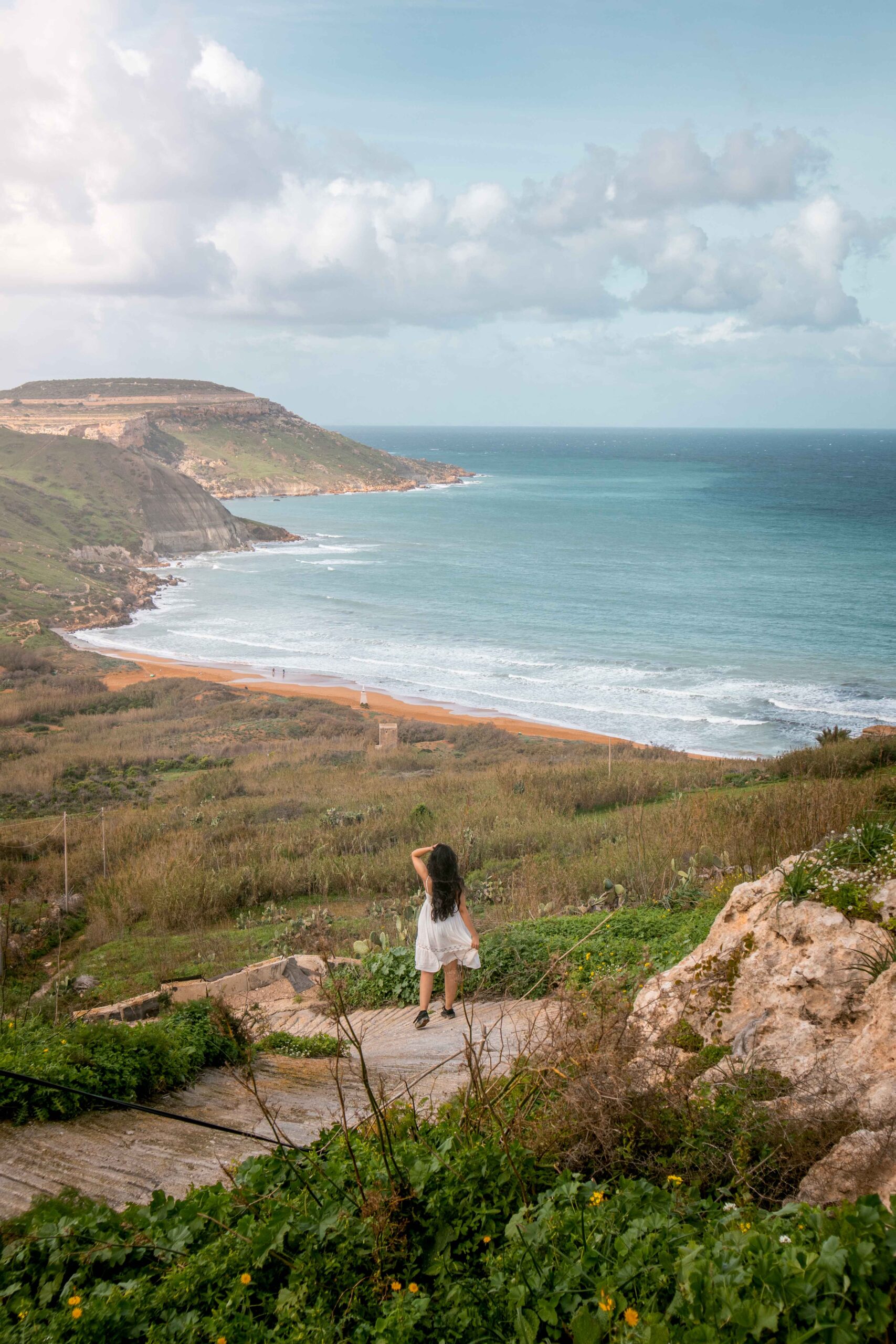Woman wearing a white dress hiking down a path to Ramla Bay Beach on Gozo island, Malta