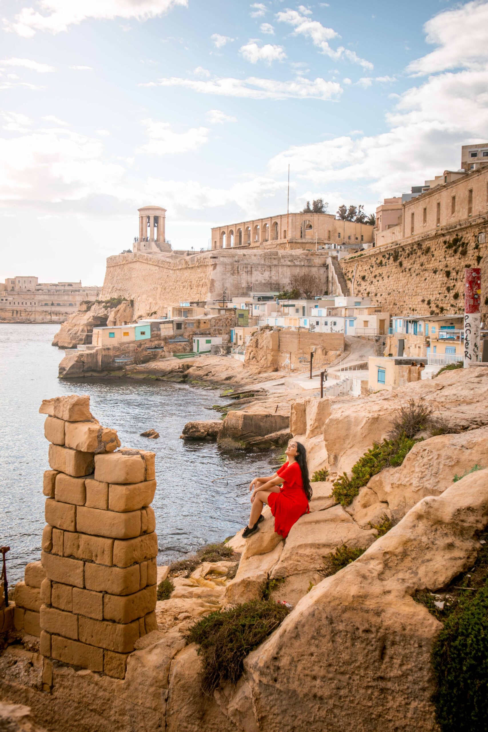 Woman wearing a red dress sitting near ruins in Wuestenwinds beach in Valletta, Malta