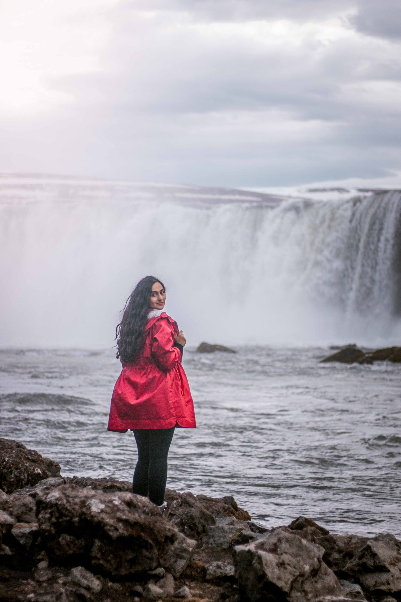 Woman wearing a red jacket posing in front of Godafoss waterfall from West bank, North Iceland