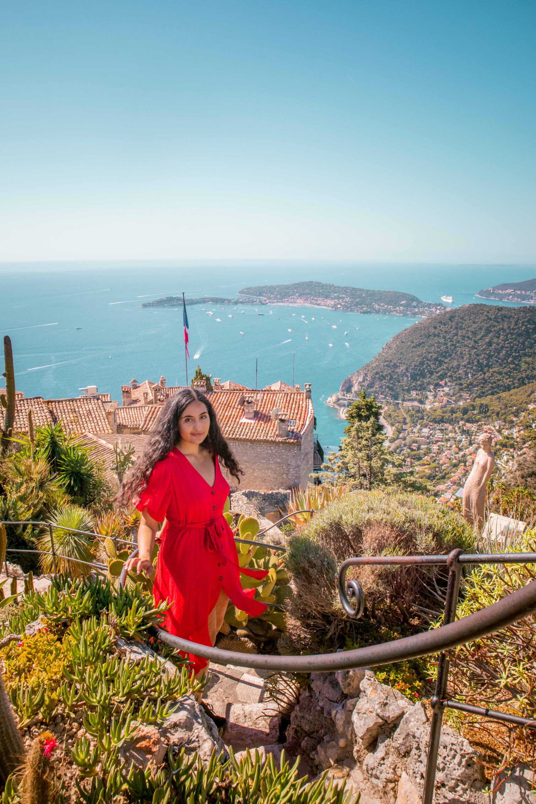 Woman wearing a red dress posing in a spiral staircase in the Eze Exotic Garden on a sunny day, featuring cacti and succulent plants in Eze Village, France