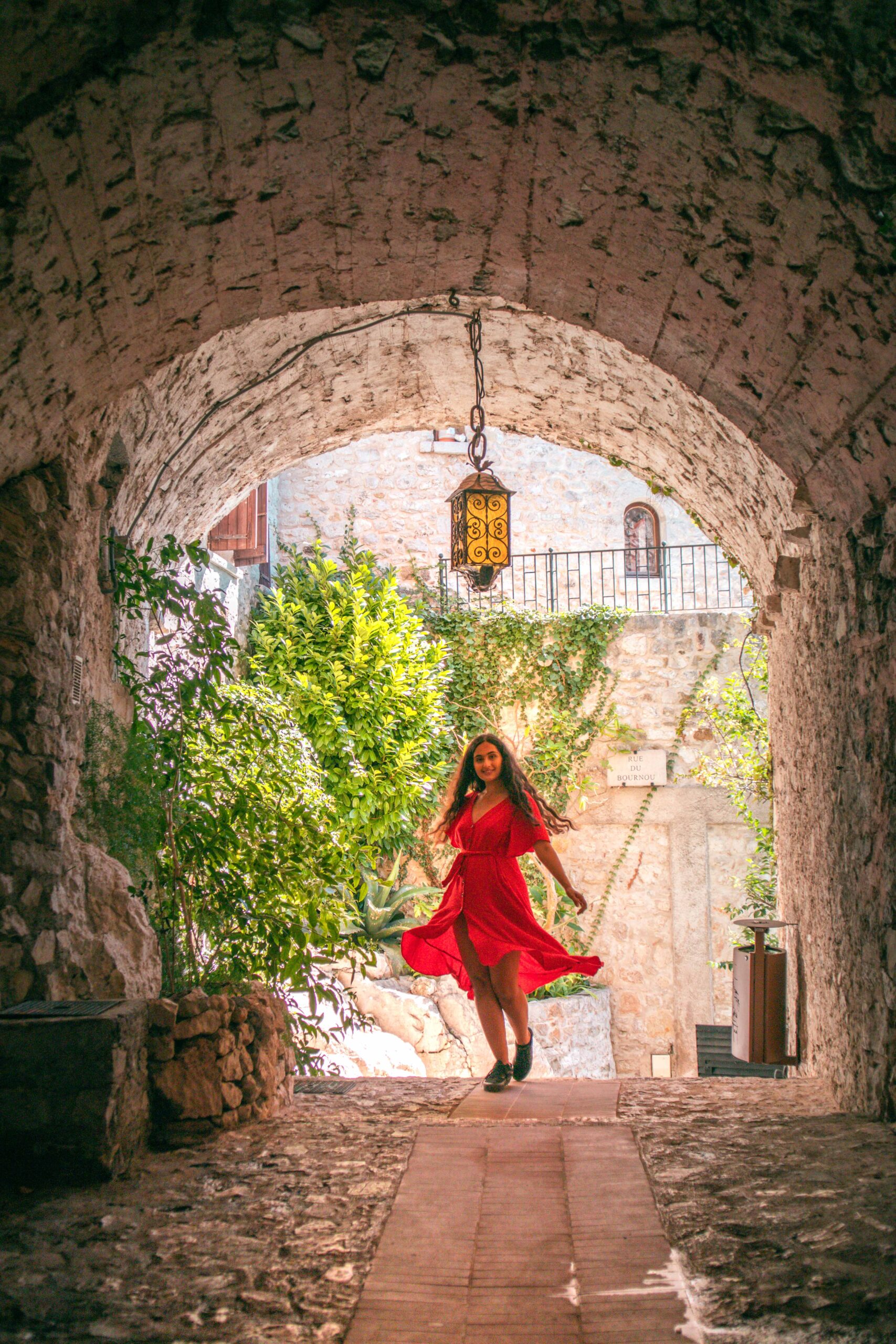 Woman wearing a red dress twirling under a stone arch near Rue du Bournou in Eze Village, France