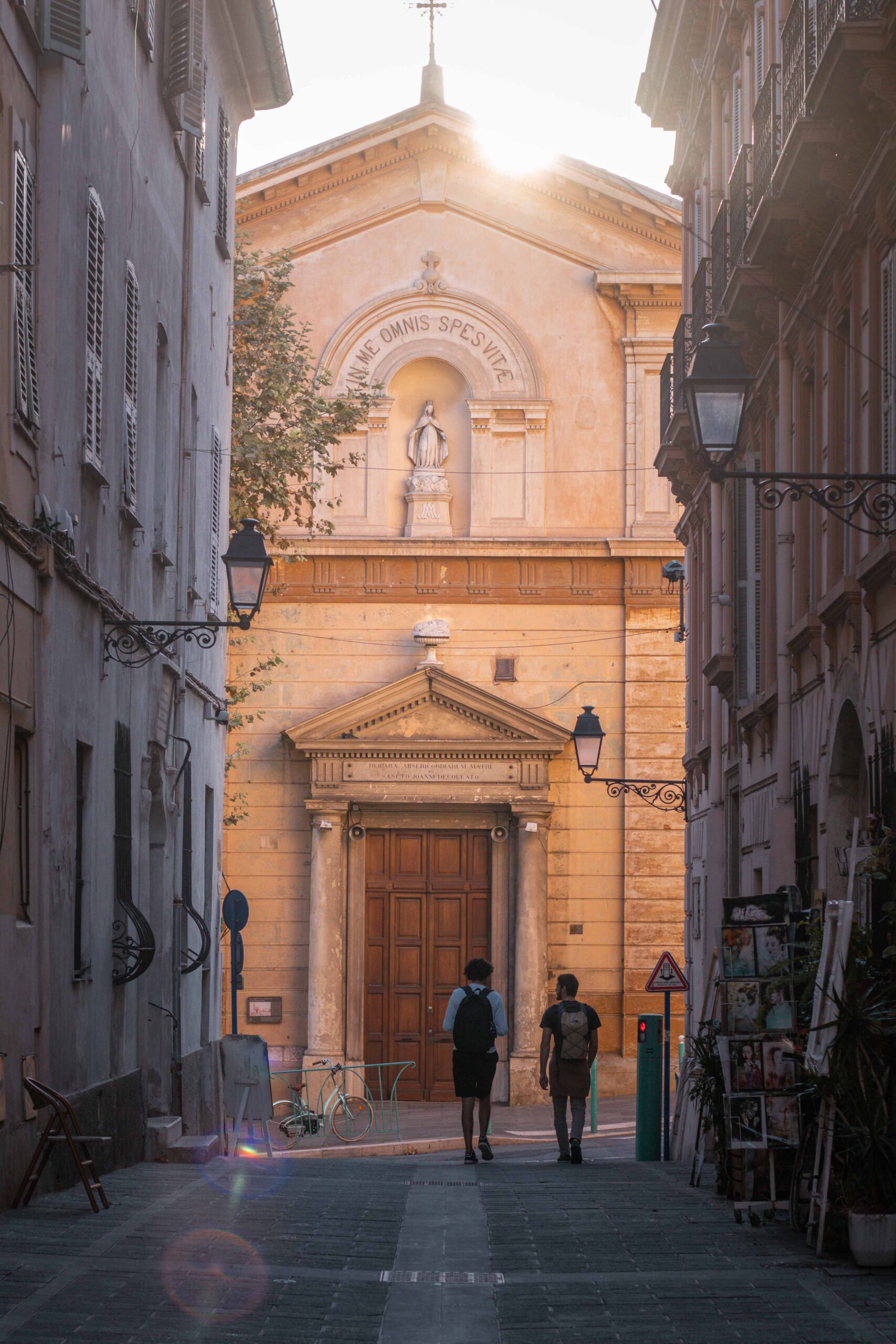 Two pedestrians walking in a street ("Rue de Bréa") towards the Chapelle de la Miséricorde in the Old Town of Menton, France