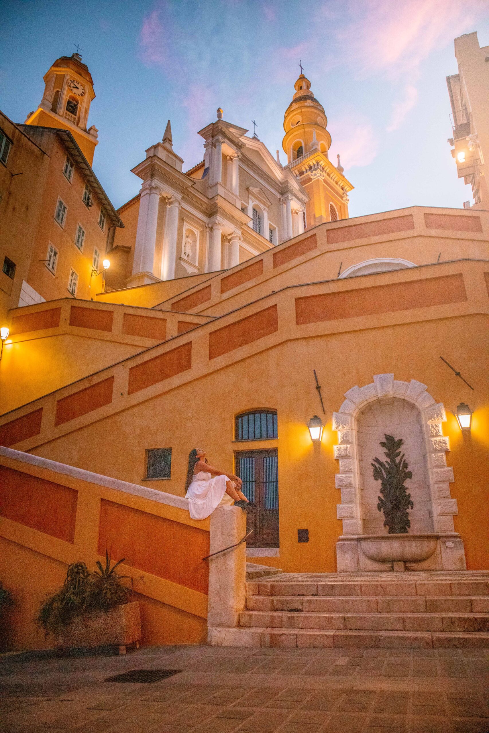 Woman wearing a white dress sitting on a yellow and orange staircase leading to the Saint Michael Archangel Basilica during sunset in the Old Town of Menton, France