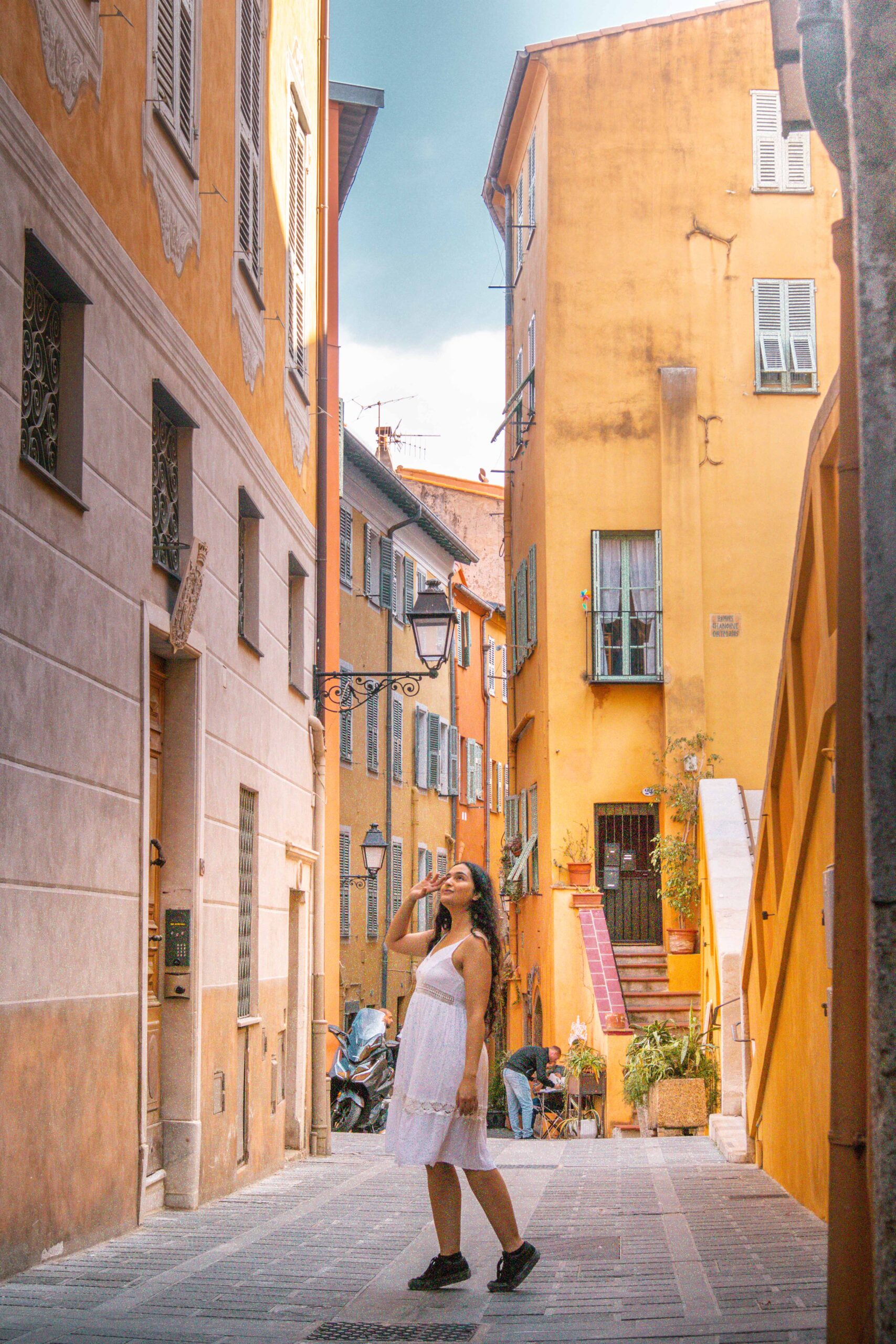 Woman wearing a white dress posing in a narrow street ("Rue Longue"), looking up at the colourful facades and windows in the Old Town of Menton, France