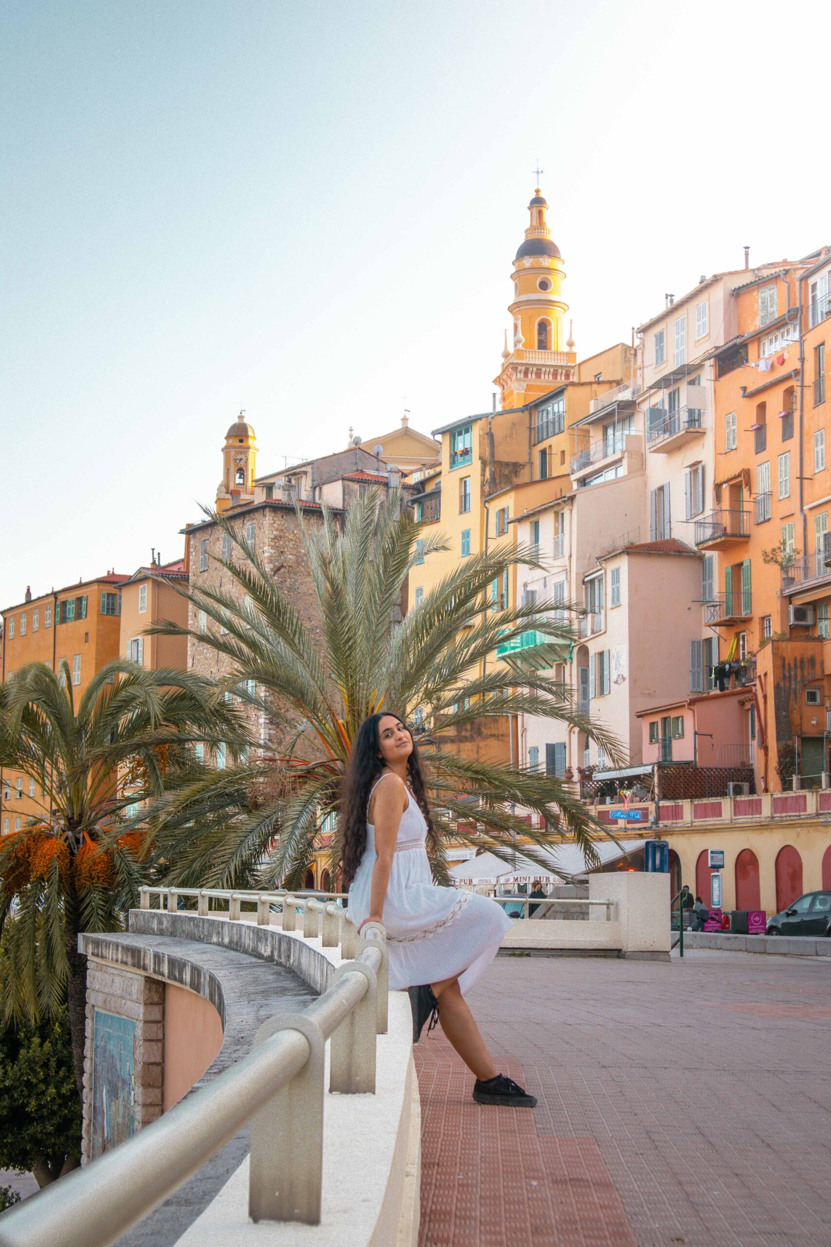 Woman wearing a white dress posing at Menton's waterfront ("Quai Bonaparte"), featuring a view of the bell tower of Saint Michael Archangel Basilica in Menton, France