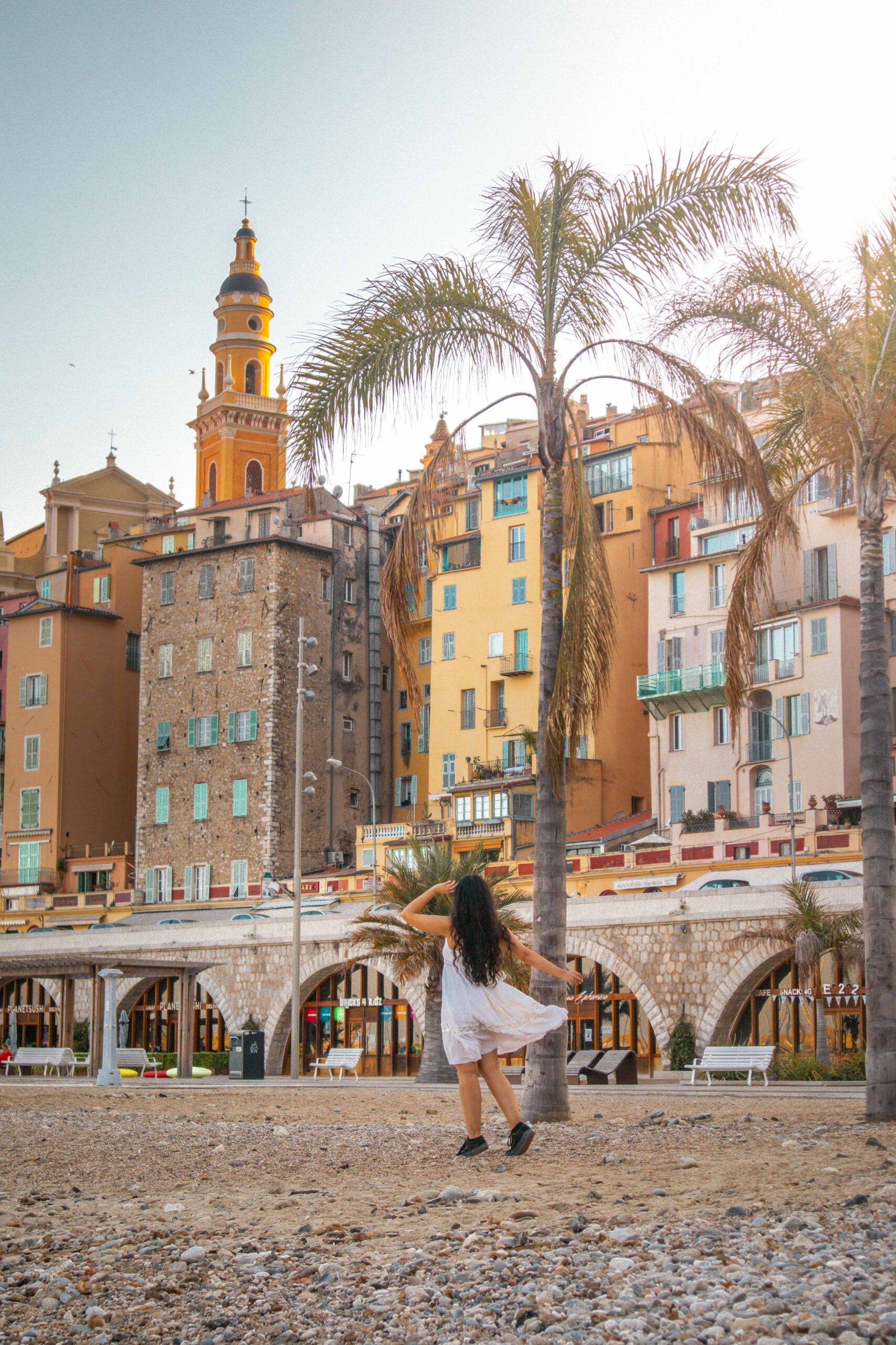 Woman wearing a white dress posing at Menton's beach, featuring a view of the bell tower of Saint Michael Archangel Basilica in Menton, France