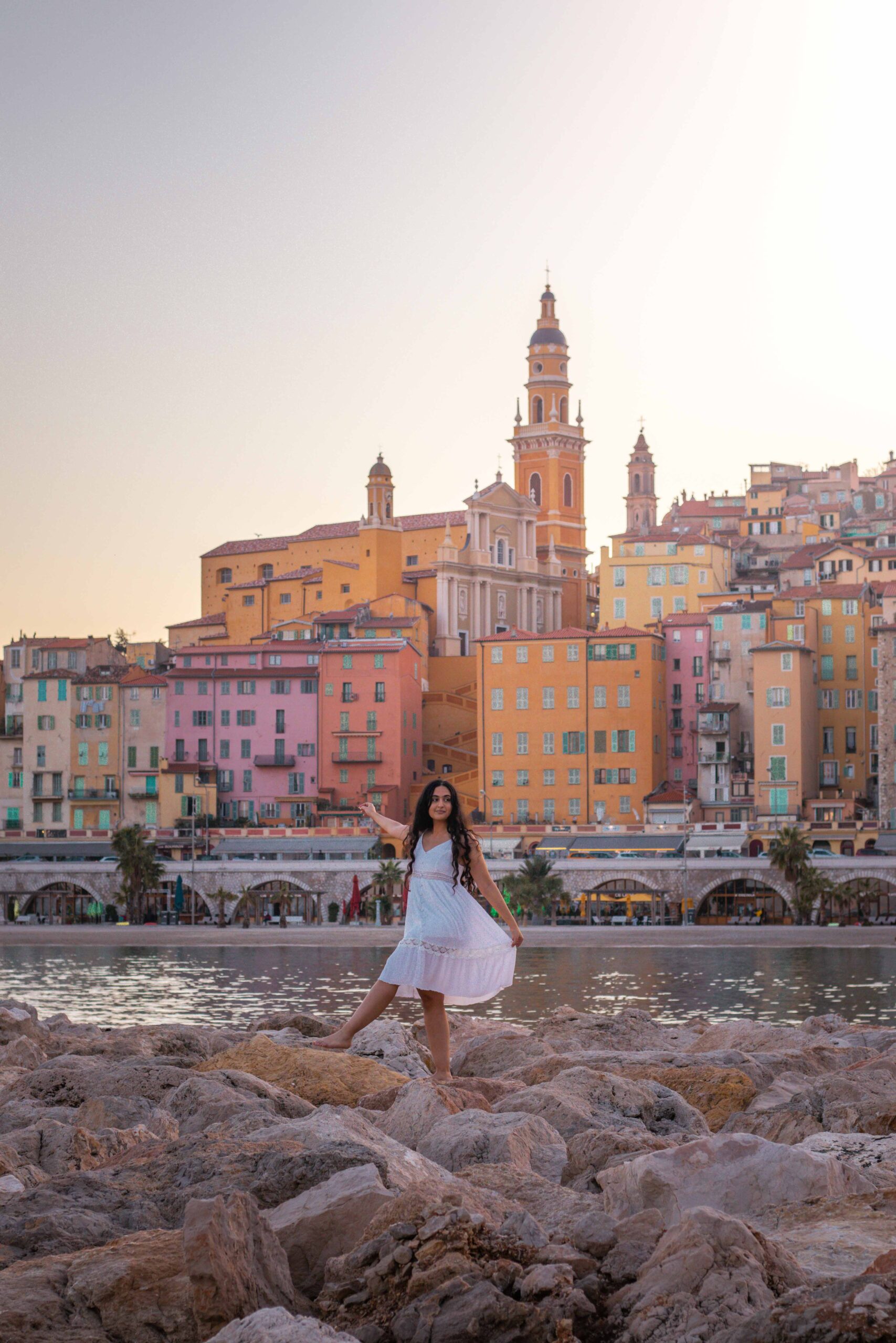 Woman wearing a white dress posing in front of Menton's beach, featuring a view of Menton's waterfront and the bell tower of Saint Michael Archangel Basilica during sunset in Menton, France