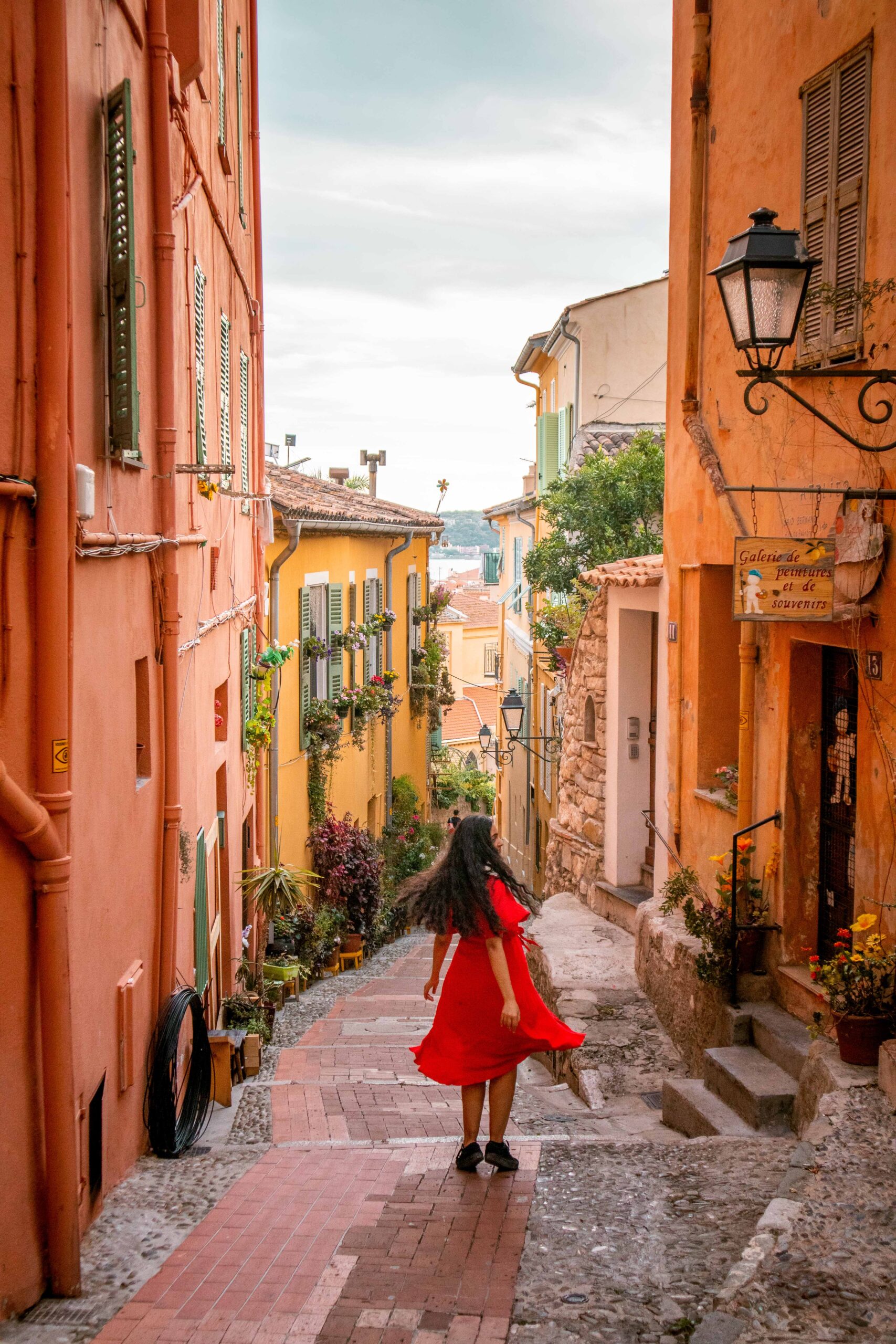 Woman wearing a red dress twirling in a colourful street ("Rue du Palmier") in front of the Saïd Berkane art gallery in the Old Town of Menton, France