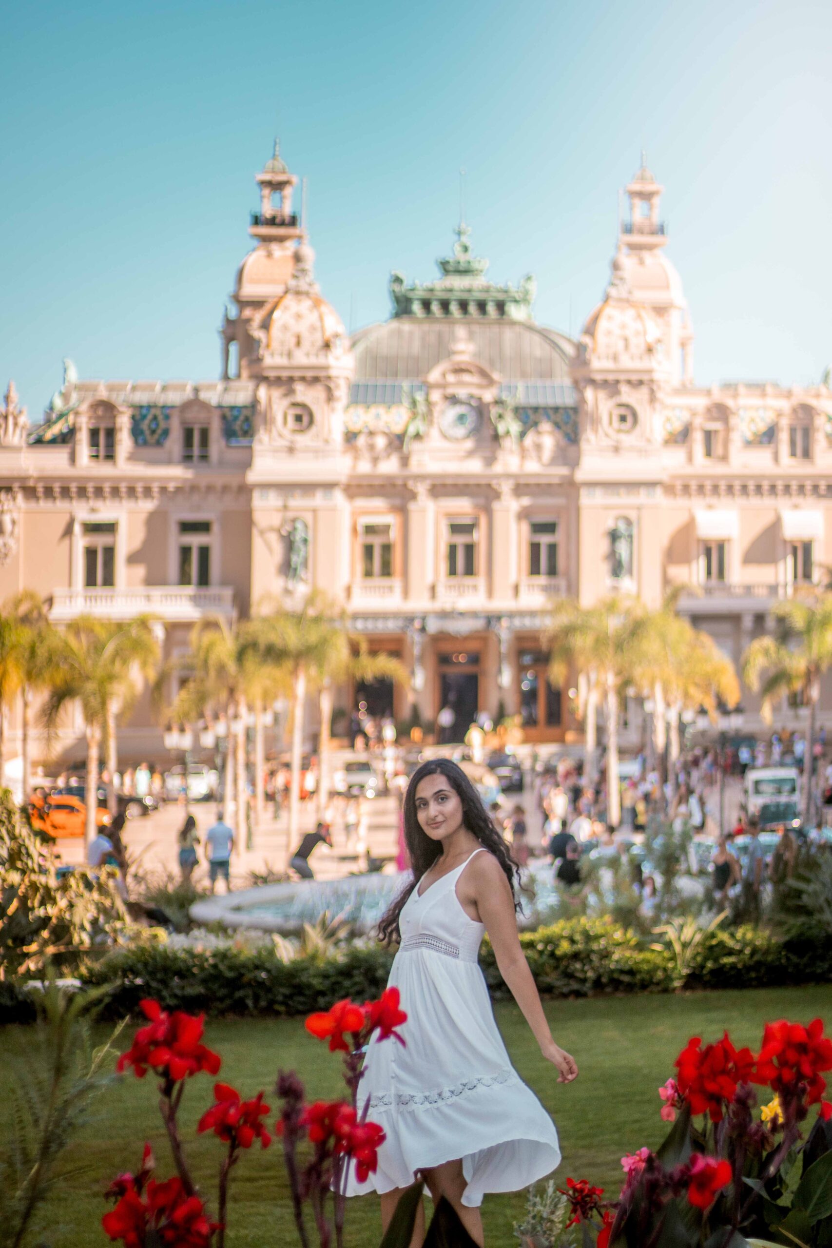 Woman wearing a white dress posing in front of the Monte-Carlo Casino in the Casino Garden during a sunny day in Monaco