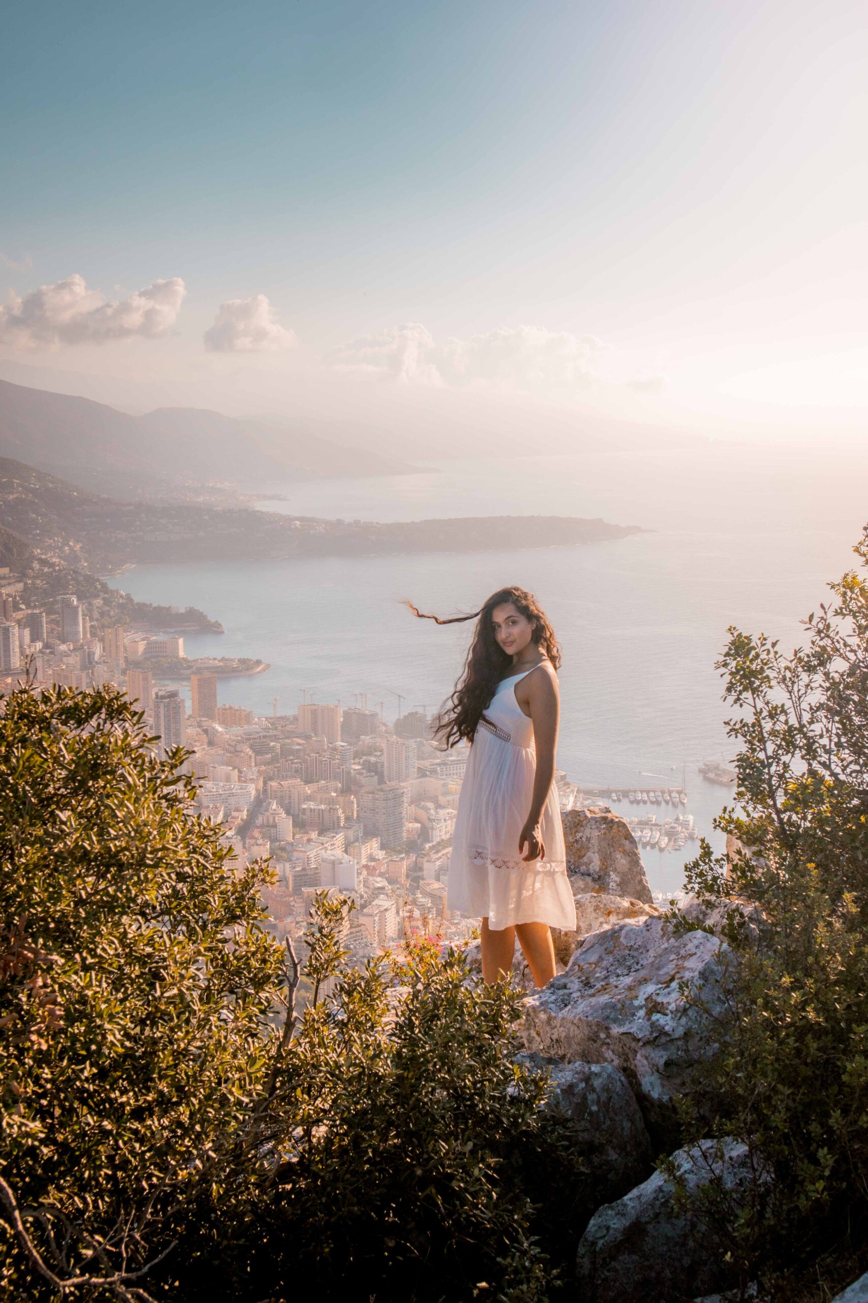 Woman wearing a white dress standing on a rock and posing in front of a panoramic view of Monaco at sunrise from the Tête de Chien rock promontory viewpoint near La Turbie Village, France