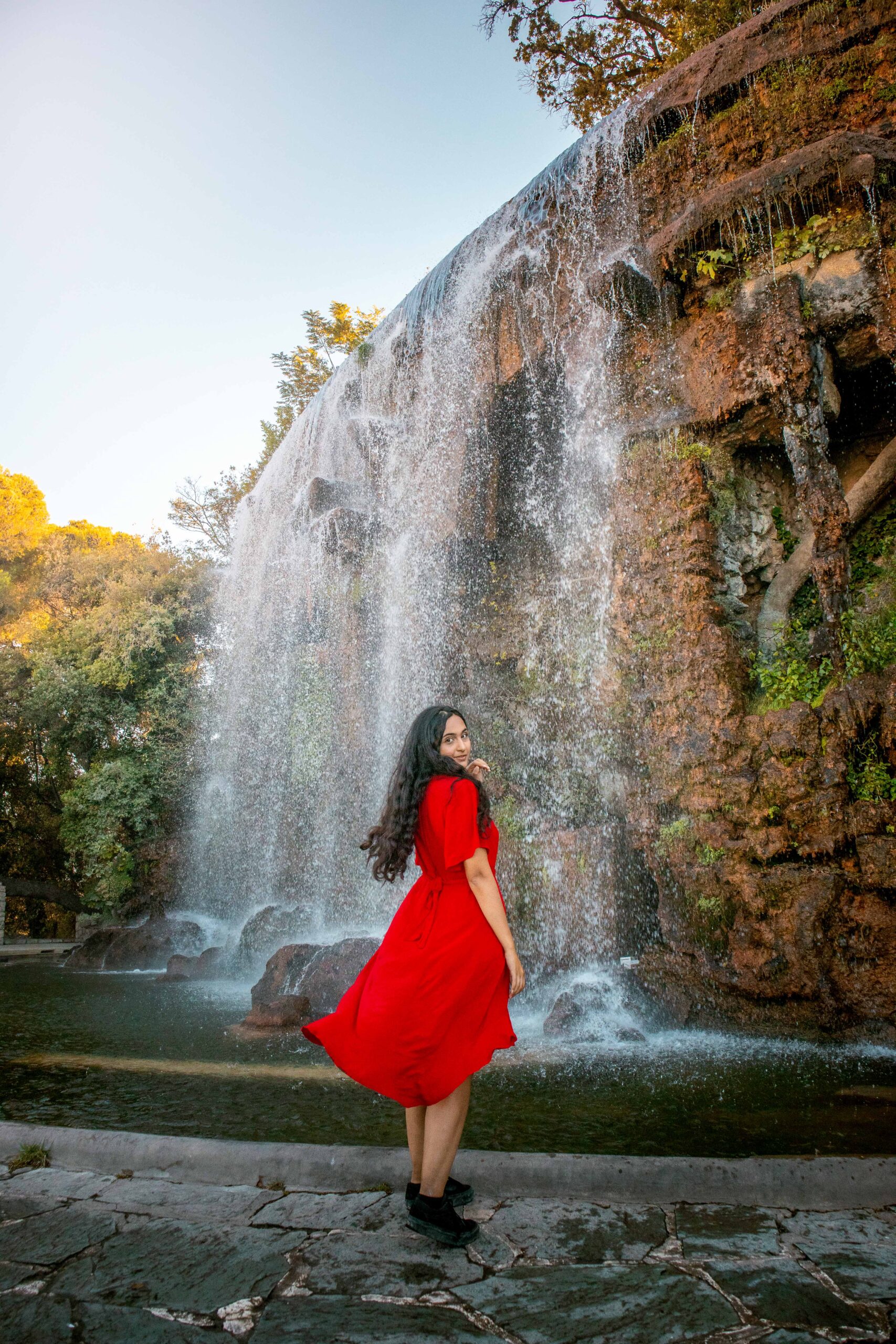 Woman wearing a red dress posing in front of the waterfall at the Colline du Château (Castle Hill) park in Nice, France