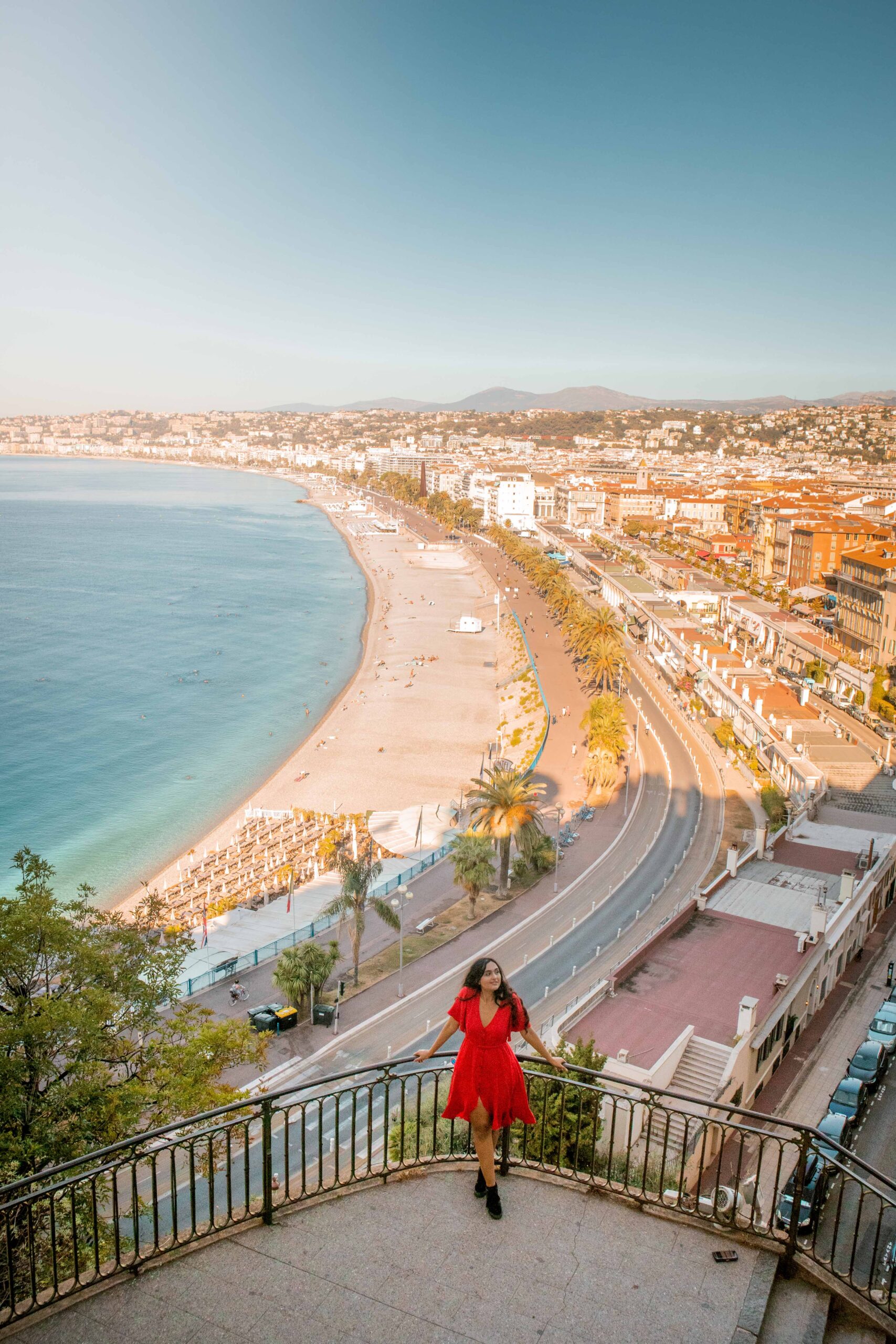 Woman wearing a red dress posing in the viewpoint balcony above the Promenade des Anglais and the pebble beach at the Colline du Château (Castle Hill) park in Nice, France