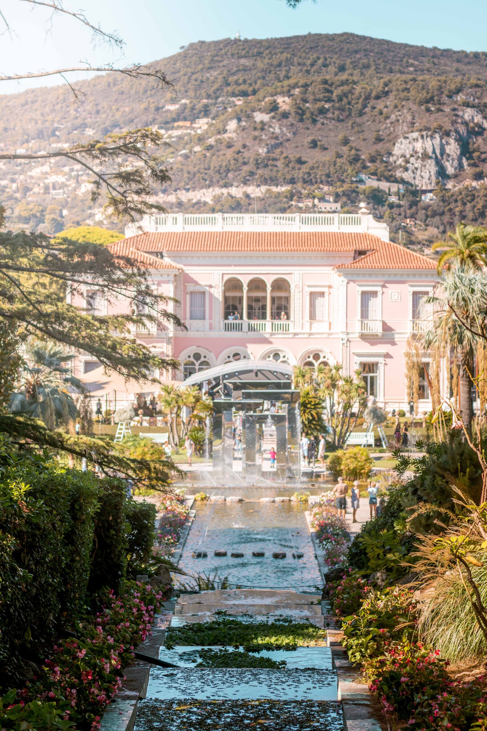 View of the pink facade of Villa Ephrussi de Rothschild as seen from the French Garden on a sunny day, featuring decorated waterfalls and ponds in Saint-Jean-Cap-Ferrat, France