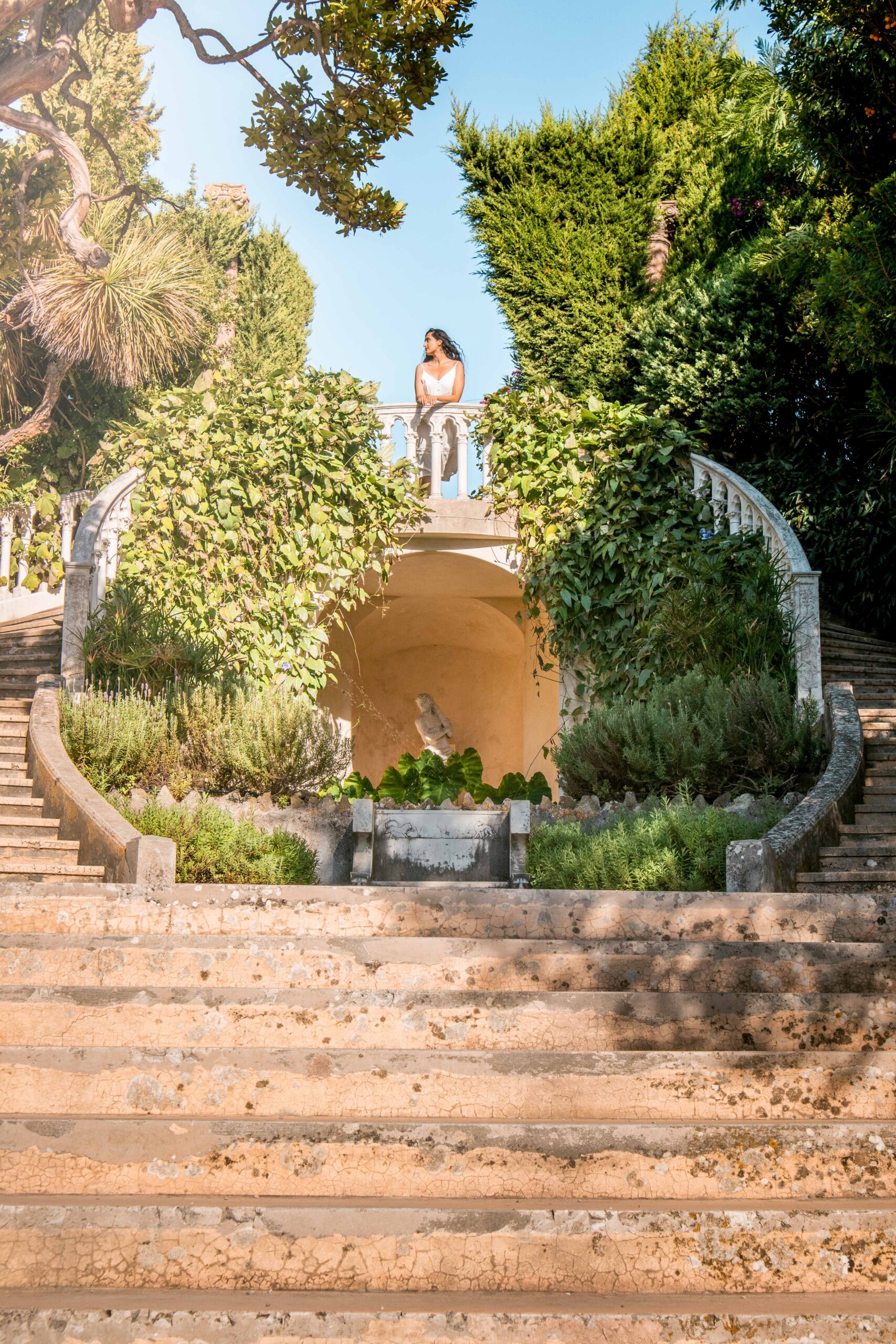 Woman wearing a white dress posing on top of the double staircase in the Florentine Garden of Villa Ephrussi de Rothschild in Saint-Jean-Cap-Ferrat, France
