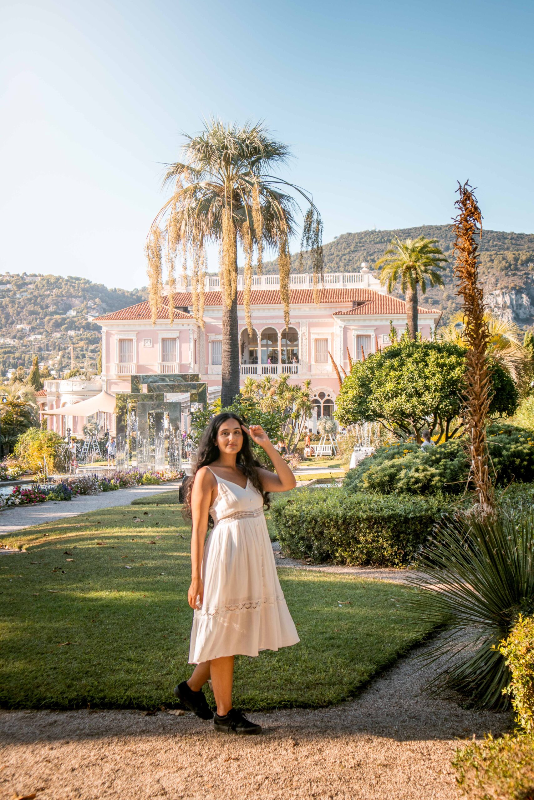 Woman wearing a white dress posing in front of the pink facade of Villa Ephrussi de Rothschild in the French Garden in Saint-Jean-Cap-Ferrat, France