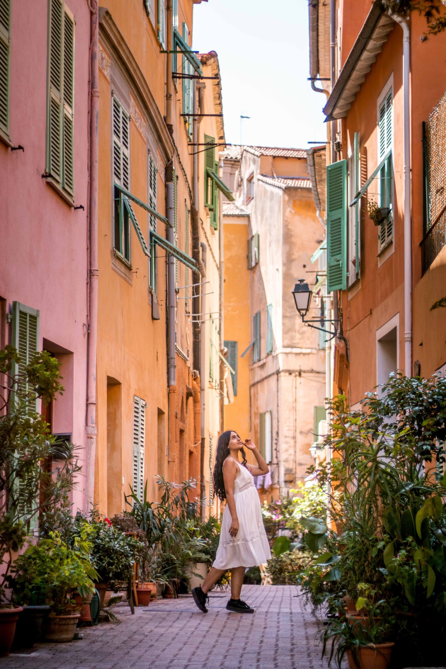 Woman wearing a white dress posing in a narrow and colourful street with several plants in the Old Town of Villefranche-sur-Mer, France
